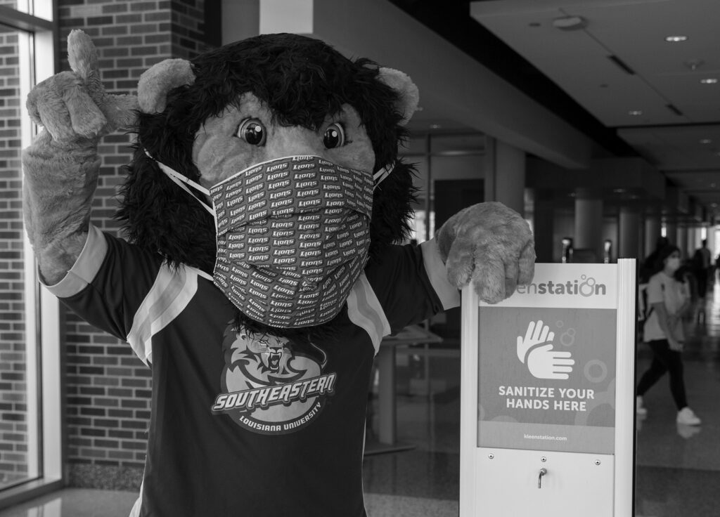 Roomie the Lion wears a mask and poses by a hand sanitation station in 2020.